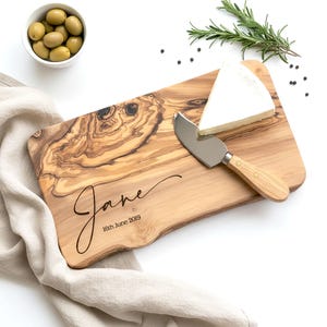 May include: A wooden cheese board with a natural wood grain pattern, featuring the name "Jane" and the date "16th June 2019" engraved in a script font. A wedge of white cheese and a cheese knife are on the board. Olives in a white bowl and rosemary sprigs are also present.