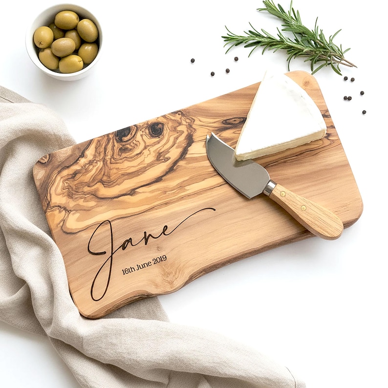 May include: A wooden cheese board with a natural wood grain pattern, featuring the name "Jane" and the date "16th June 2019" engraved in a script font. A wedge of white cheese and a cheese knife are on the board. Olives in a white bowl and rosemary sprigs are also present.