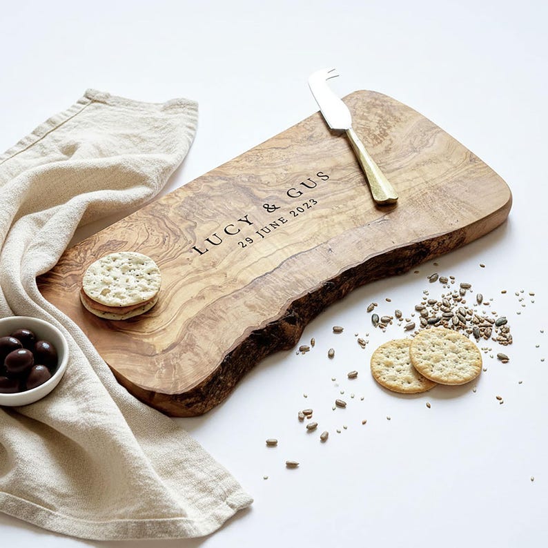 May include: A wooden serving board with a natural edge, engraved with "LUCY & GUS 29 JUNE 2023". A cheese knife rests on the board. Crackers, olives, and a linen napkin are arranged around the board.