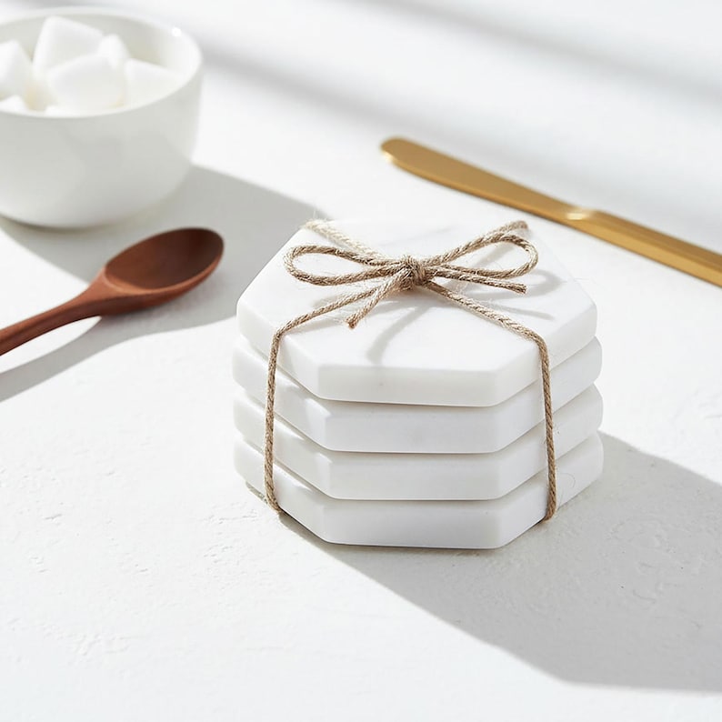 May include: A stack of four white marble hexagonal coasters tied with twine. A wooden spoon, a small white bowl with sugar cubes, and a gold butter knife are also in the image, all on a white surface.