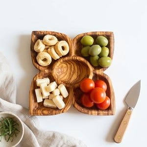 May include: A wooden serving platter with four sections, containing snacks. The platter holds cheese cubes, green olives, cherry tomatoes, and small bread rings. A small knife with a wooden handle is placed next to the platter.
