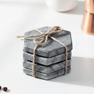 May include: A stack of four gray hexagonal coasters tied with twine. The coasters have a marbled appearance and are made of a stone-like material. The twine is tied in a bow, suggesting a gift or set. The background is white.
