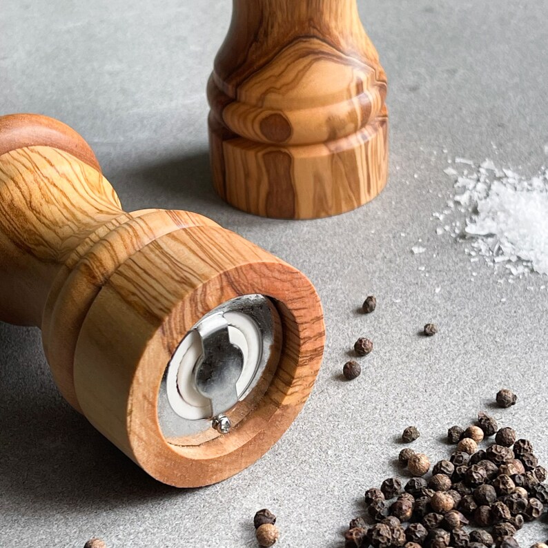 May include: Two wooden pepper mills with a natural wood grain finish. One mill is in the foreground, showing the grinding mechanism. Scattered around are peppercorns and salt crystals, suggesting culinary use.