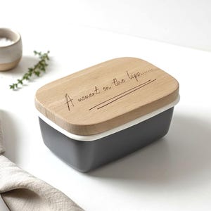May include: A rectangular butter dish with a wooden lid. The lid is engraved with the phrase "A moment on the lips..." in a handwritten style. The dish is dark gray with a white rim.