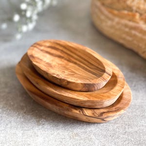 May include: A stack of three oval-shaped wooden dishes. The dishes are made of light brown wood with a visible grain pattern. The smallest dish sits on top of the other two, creating a layered effect. The dishes are on a gray surface.