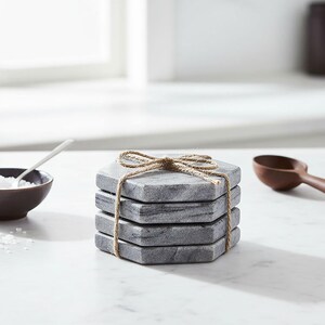 May include: A stack of four gray marble coasters tied with twine. The coasters are hexagonal and have a natural stone appearance. A small bowl with salt and a wooden spoon are in the background.