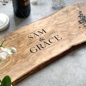 May include: A wooden serving board with a live edge. The board is engraved with the text "SAM & GRACE EST. 1999" in black. The wood has a natural grain and a light brown colour. The board is ideal for serving food.