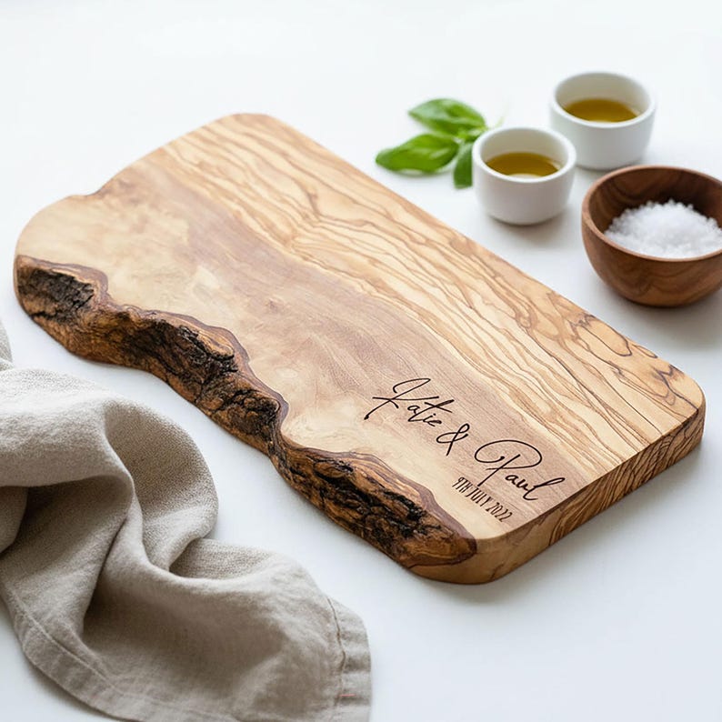 May include: A wooden cutting board with a live edge, engraved with "Katie & Paul" and the date "9th July 2022". The board is light brown with dark brown accents. Small bowls of oil and salt are nearby.