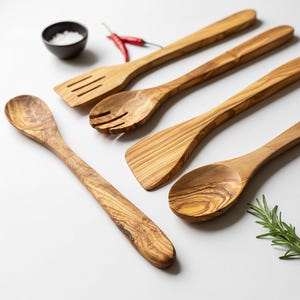May include: A collection of wooden kitchen utensils, including a slotted spoon, spatula, and serving spoons. The utensils are made of light-colored wood with a natural grain pattern. A small bowl of salt, red peppers, and rosemary sprig are also present.