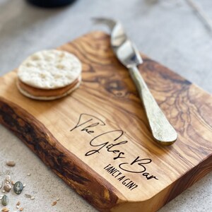 May include: A wooden serving board with a natural edge, featuring the text "The Giles Bar Fancy a Gin?" A cheese knife rests on the board next to a cracker sandwich. The board has a light brown color with darker wood grain.