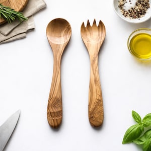 May include: Wooden salad spoon and fork set. The spoon has a rounded bowl, and the fork has tines. The wood grain is visible. The set is next to a small bowl of salt and pepper, a bowl of olive oil, and fresh herbs.