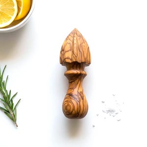 May include: A wooden citrus reamer with a conical head and a rounded handle. The reamer is made of light brown wood with a visible grain pattern. The image also includes a bowl of lemon slices, rosemary sprigs, and salt.