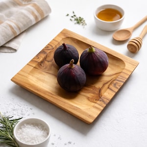 May include: Three ripe figs with dark purple skin sit on a square wooden serving tray. The tray is made of light-coloured wood with a natural grain. Small bowls of honey and salt, along with wooden spoons, are also present, suggesting a culinary theme.