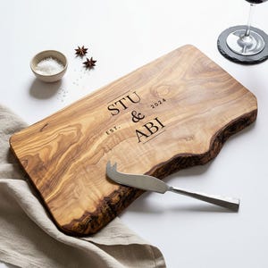May include: A wooden serving board with a natural edge, engraved with "STU & ABI EST. 2024". A cheese knife rests on the board. A small bowl of salt and star anise are nearby. The board is made of light brown wood.