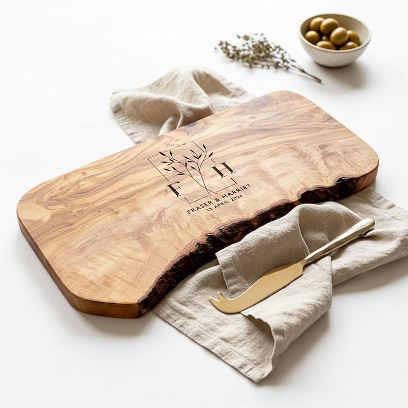 May include: A rectangular wooden serving board with a live edge, featuring engraved initials and a date. The board is light brown with a natural wood grain. A gold cheese knife rests on a beige linen napkin. A small bowl of olives is in the background.