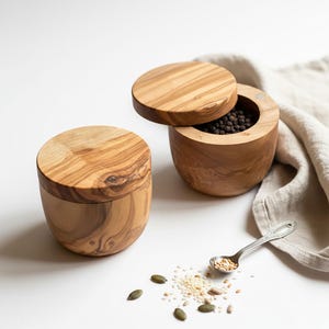 May include: Two wooden salt and pepper cellars with lids, one open to reveal peppercorns. A small spoon holds seeds, with scattered seeds and a linen cloth in the background. The cellars are made of light-coloured wood.