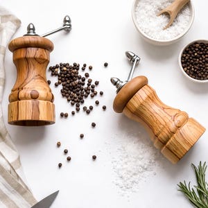 May include: Two wooden pepper grinders with stainless steel handles and tops are displayed on a white surface. Scattered peppercorns and bowls of salt and peppercorns are also visible. A striped cloth and a knife are in the background.