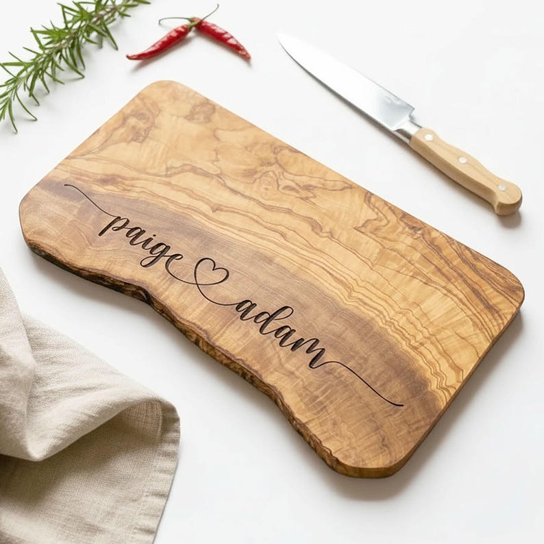 May include: A wooden cutting board with a natural edge, engraved with the names "paige" and "adam" separated by a heart symbol. A stainless steel knife with a light-colored wooden handle rests on the side. Red chili peppers and rosemary sprigs are also present.