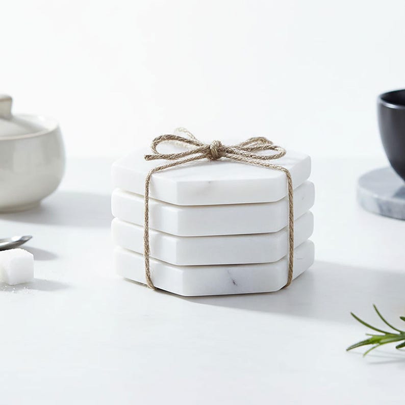 May include: A stack of four white marble hexagonal coasters tied with twine. The coasters have a polished surface and are stacked neatly. The twine is tied in a bow at the top, adding a decorative touch.