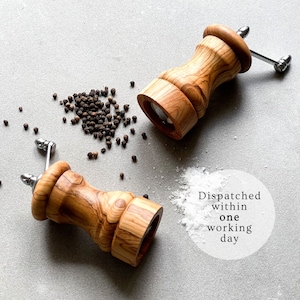 May include: Two wooden pepper grinders with stainless steel handles and a scattering of black peppercorns and white salt on a gray surface. The grinders have a light brown wood grain.