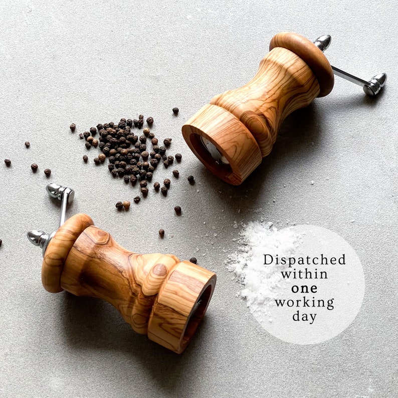 May include: Two wooden pepper grinders with stainless steel handles and a scattering of black peppercorns and white salt on a gray surface. The grinders have a light brown wood grain.