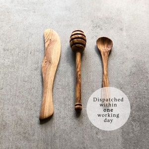 May include: Three wooden kitchen utensils: a butter knife, a honey dipper, and a spoon. All three utensils are made of olive wood and have a natural wood finish.