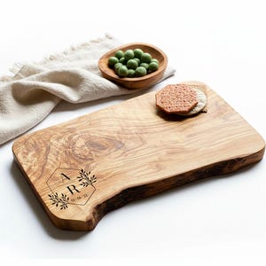 May include: A wooden serving board with a natural edge, engraved with the letters "A R" and the date "13-06-22" within a decorative frame. The board holds crackers and sits near a small wooden bowl with green snacks and a beige linen napkin.