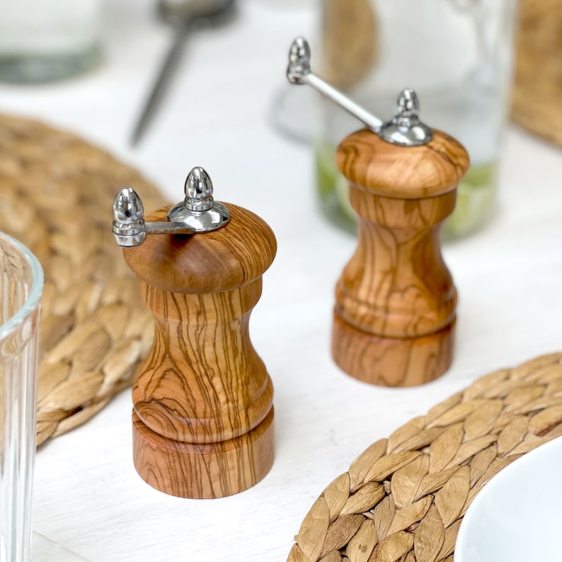 May include: Two wooden pepper grinders with a natural wood grain finish and silver-colored metal tops and handles. The grinders are on a white surface with woven placemats and glassware in the background. A kitchen or dining room accessory.