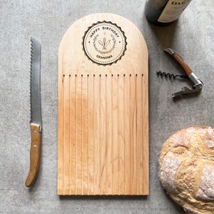 Pu&ograve; includere: Un tagliere di legno chiaro con un coltello seghettato, una bottiglia di vino e un cavatappi. Il tagliere ha incise le parole "Happy Birthday Grandma".