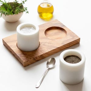 May include: A wooden serving tray with a circular indentation and two white marble salt and pepper cellars. A small silver spoon rests on the surface. A small bowl of herbs and a bottle of oil are in the background.