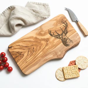 May include: A wooden serving board with a deer head design, a cheese knife, a linen napkin, cherry tomatoes, and crackers. The board has a natural wood grain pattern and a unique shape. The cheese knife has a wooden handle.