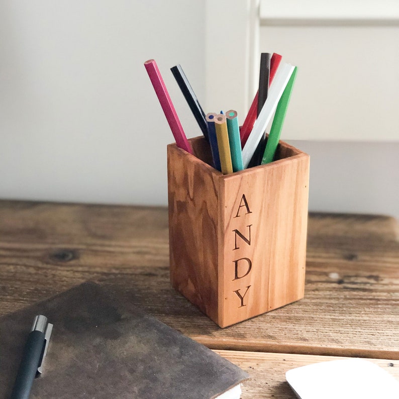 May include: A wooden pencil holder with the name "ANDY" engraved on the front. The holder is filled with colored pencils, including red, green, blue, and yellow.