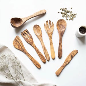 May include: A collection of handcrafted olive wood kitchen utensils, including a ladle, slotted spatula, salad fork, spoon, and butter knife. The wooden tools display a natural grain pattern and are arranged on a white surface, suggesting a rustic aesthetic.