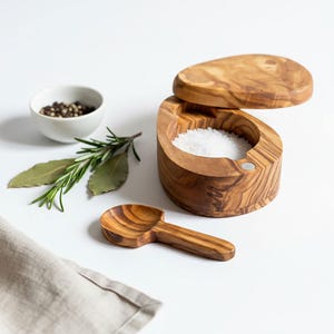 May include: A wooden salt pig with a matching lid and a small wooden spoon. The salt pig is filled with white salt. A small white bowl of peppercorns, rosemary, and bay leaves are also present.