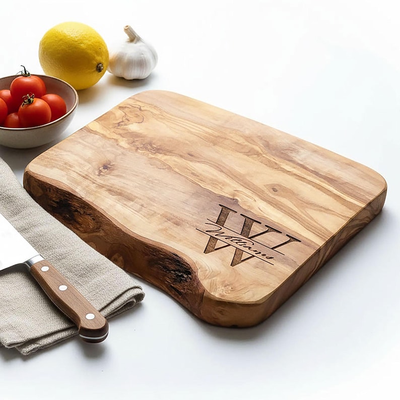 May include: A rectangular wooden cutting board with a natural edge. The board has a light brown color with darker wood grain patterns. The initials "IV" and the name "Williams" are engraved on the board. A knife and vegetables are in the background.