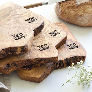 May include: A stack of wooden cutting boards with natural edges and the "Yeo Valley" logo. The boards are light brown with darker brown edges. A loaf of bread and a knife are in the background. A sprig of white flowers is in the foreground.