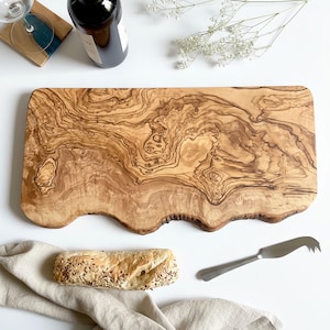May include: A large, rectangular wooden cutting board with a wavy edge. The board has a natural wood grain pattern and is surrounded by a baguette, cheese knife, wine bottle, and a wine glass.