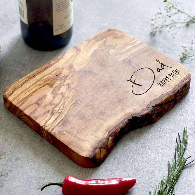 May include: A wooden cutting board with a natural edge, featuring the word "Dad" and "Happy 90th!" engraved on the surface. The board has a light brown color with darker wood grain patterns. A red chili pepper and rosemary sprig are in the foreground.