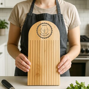 Pu&ograve; includere: Una lama per pane in legno chiaro con la parte superiore arrotondata e scanalature verticali. La parte superiore presenta un emblema circolare con il testo "Gran's Delicious Cakes!" e illustrazioni di utensili da forno. La lama per pane &egrave; tenuta da una persona con un grembiule nero.