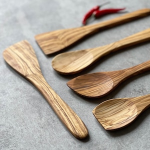 May include: Five wooden kitchen utensils, including spoons and spatulas, made from olive wood with a natural wood grain pattern. The utensils are arranged on a gray surface.