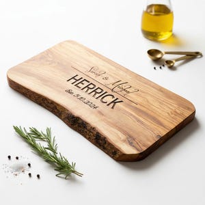May include: A rectangular wooden cutting board with a live edge. The board is engraved with the names "Sarah & Michael" and "HERRICK", along with the date "Est. 5.10.2024". Rosemary, salt, pepper, and oil are also present.