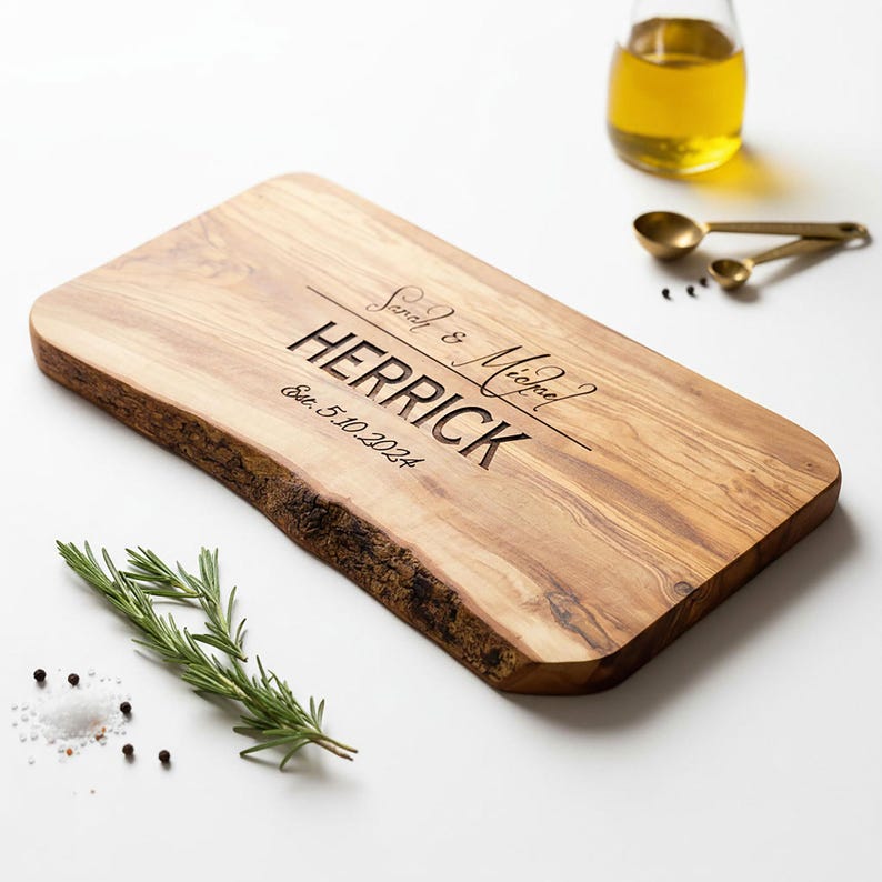 May include: A rectangular wooden cutting board with a live edge. The board is engraved with the names "Sarah & Michael" and "HERRICK", along with the date "Est. 5.10.2024". Rosemary, salt, pepper, and oil are also present.