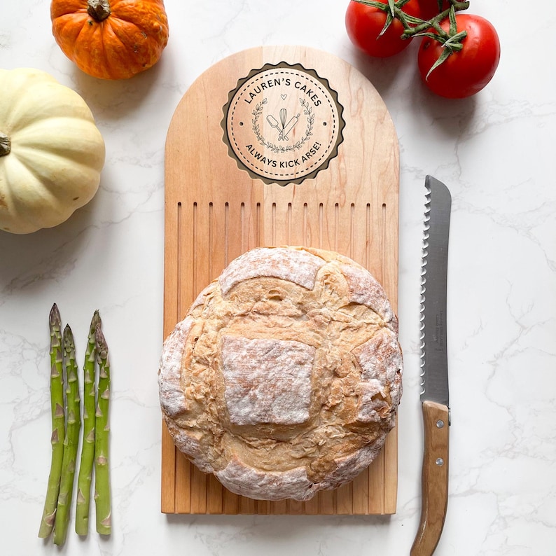 Pu&ograve; includere: Un tagliere di legno con una pagnotta di pane, un coltello seghettato e la scritta "LAUREN'S CAKES ALWAYS KICK ARSE!". La tavola ha un design decorativo ed &egrave; circondata da verdure, tra cui pomodori, asparagi e zucche.