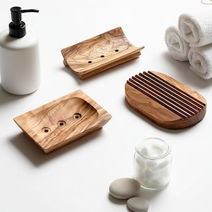 May include: A collection of wooden soap dishes and a white ceramic soap dispenser. The soap dishes are made of olive wood, showcasing a natural grain pattern. One dish has a slatted design, while others have drainage holes. A glass jar with cotton balls is also present.