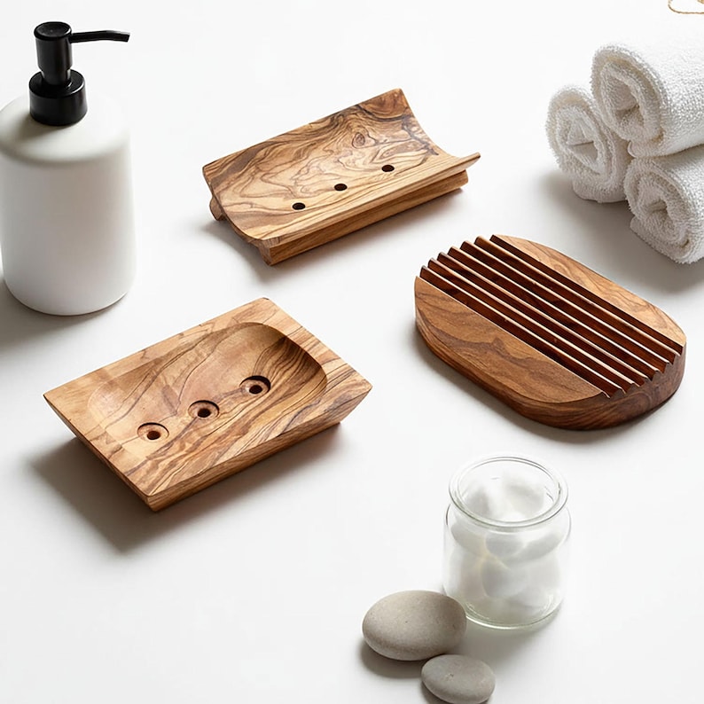 May include: A collection of wooden soap dishes and a white ceramic soap dispenser. The soap dishes are made of olive wood, showcasing a natural grain pattern. One dish has a slatted design, while others have drainage holes. A glass jar with cotton balls is also present.