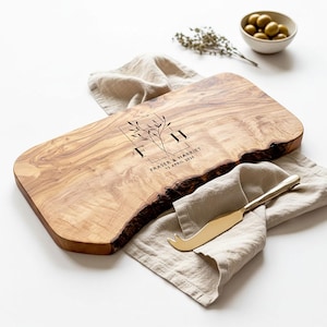 May include: A wooden serving board with a live edge, featuring engraved initials and the text "Fraser & Harriet 13 April 2024". A gold cheese knife rests on a beige linen napkin. A small bowl of olives is in the background.