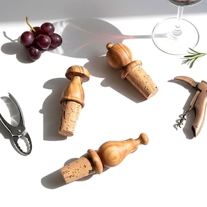 May include: Wooden wine bottle stoppers with cork bases, a wine opener, grapes, and a wine glass. The stoppers have distinct shapes and are made of light-coloured wood. The items are arranged on a white surface, creating a still-life composition.