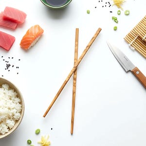 May include: Overhead shot of sushi ingredients on a white surface. Includes sushi, rice, soy sauce, chopsticks, a knife, and garnishes. The chopsticks are made of wood. The sushi includes salmon and tuna.