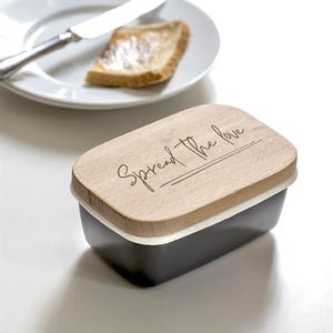 May include: A black rectangular butter dish with a wooden lid. The lid is engraved with the words "Spread the love". A white rim separates the lid from the black base. A plate with a knife and a slice of toast is in the background.
