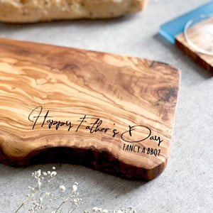 May include: A wooden cutting board with a natural edge, featuring the text "Happy Father's Day" and "Fancy a BBQ?" in black script. The board has a light brown color with darker wood grain patterns.